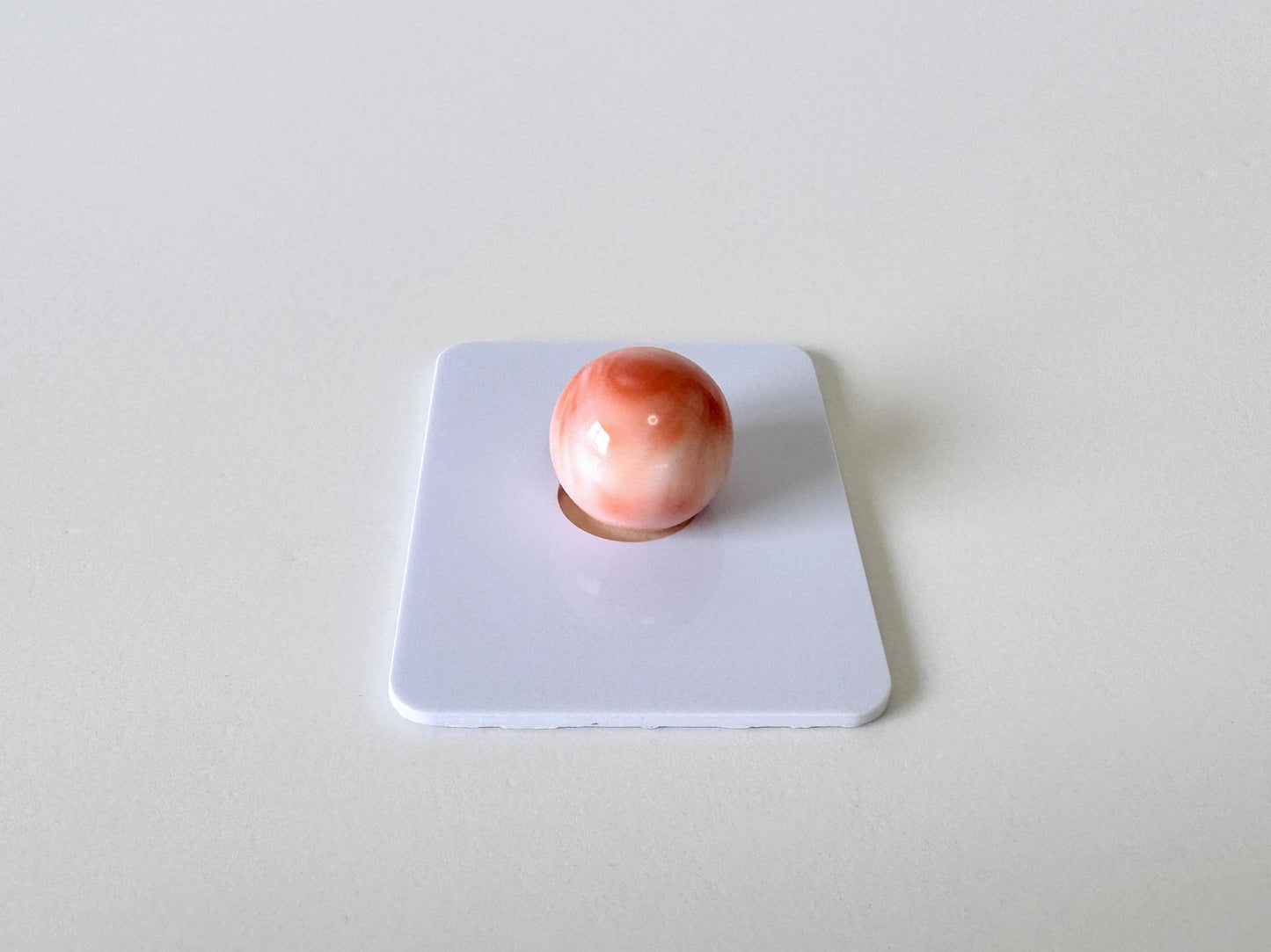 A single, round, pink and white marble rests on a white square surface.