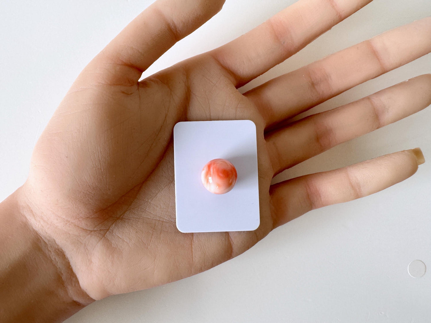 A hand holding a small white square with a red and orange marble in the center.