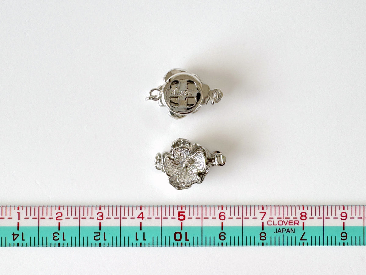 two silver-colored flower-shaped charms with a cross design, placed next to a red and blue measuring tape.